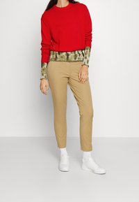 Nainen, joka pitää yllä punaista villapuseroa vihreän sideharsopaitansa päällä, beiget housut, valkoiset sukat ja valkoiset tennarit, seisoo tasaista taustaa vasten.