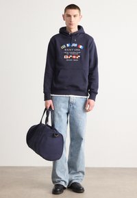 Felpa con cappuccio blu navy con stampa grafica e tasca frontale, abbinata a jeans azzurri e a una borsa da viaggio blu navy coordinata. Scarpe nere.