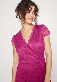 Vestido de renda fúcsia com mangas curtas e acabamento ondulado e decote em V. Apresenta padrões florais e uma silhueta ajustada. Textura suave.