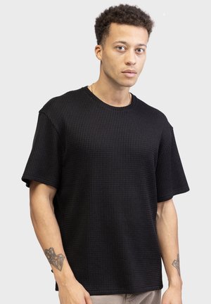 Black waffle-textured short-sleeve t-shirt with a crew neckline and relaxed fit, displayed on a male model against a gray background.