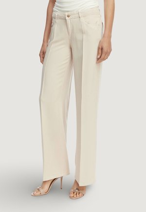 Donna che indossa pantaloni beige chiaro a gamba larga e sandali beige con tacco alto con dettagli decorativi in metallo sulle dita dei piedi.
