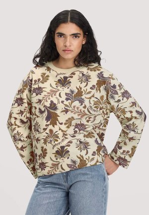Jonge vrouw met zwart krullend haar draagt een beige trui met paarse en bruine bloemmotieven en een blauwe spijkerbroek, staand met één hand op de heup.