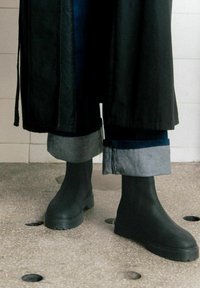 Botas de tobillo de goma negras con una suela gruesa, combinadas con unos jeans de mezclilla oscuros arremangados; un abrigo largo negro cae sobre el atuendo.