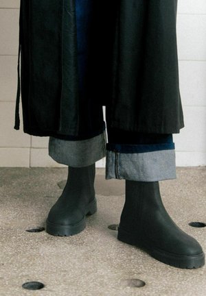 Stivali alla caviglia in gomma nera con suola spessa, abbinati a jeans scuri di denim arrotolati; un lungo cappotto nero scivola sull'outfit.
