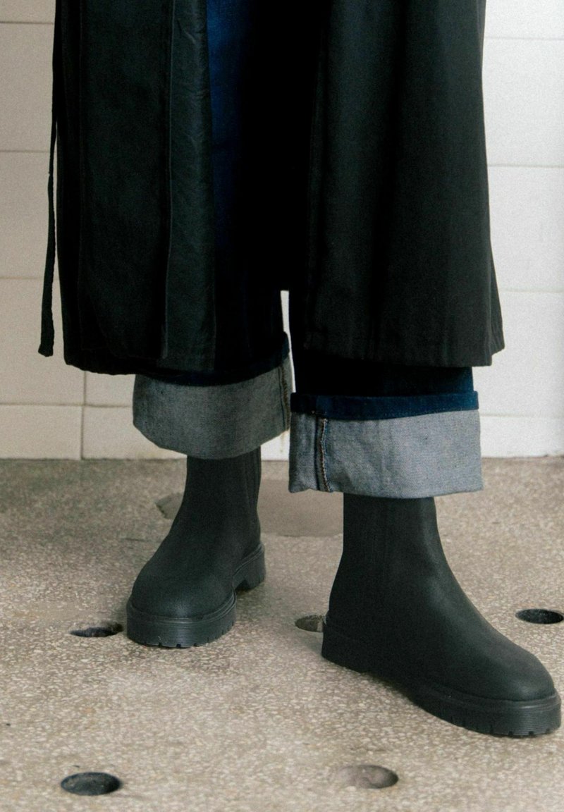Botas de tobillo de goma negras con una suela gruesa, combinadas con unos jeans de mezclilla oscuros arremangados; un abrigo largo negro cae sobre el atuendo.