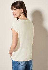 Cremefarvet T-shirt med korte ærmer og afslappet pasform samt en subtil tekstur, parret med blå denim jeans. Set fra bagsiden.