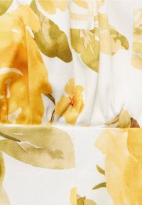 Gros plan sur un tissu à imprimé floral jaune et vert sur fond blanc, montrant des plis et un détail de couture.