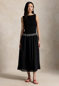 Nainen pukeutuneena mustaan hihattomaan mekkoon, jossa valkoiset geometriset reunukset vyötäröllä, seisomassa neutraalilla taustalla, näkyvissä punaiset sandaalit.