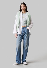 Biała koszula z guzikami noszona na jasnej zielonej bluzie, w połączeniu z jasnoniebieskimi dżinsami o wysokim stanie i białymi trampkami. Prosty, casualowy design.
