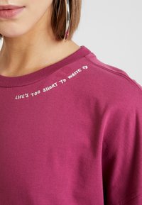 Kurzarm-T-Shirt mit Rundhalsausschnitt aus tiefpurpurfarbenem Stoff mit weißem gesticktem Text auf der Schulter, der "DAS LEBEN IST ZU KURZ, UM ES ZU VERSCHWENDEN" liest.