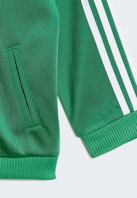 Zielona sportowa kurtka wykonana z fakturowanej tkaniny, z białymi paskami, ściągaczami na mankietach oraz boczną kieszenią z ukrytym zamkiem.