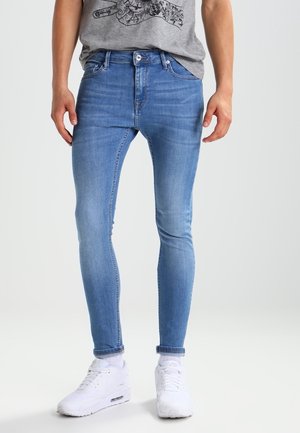 Mann trägt hellblaue Skinny-Jeans, graues T-Shirt mit grafischem Handmotiv, weiße Socken und weiße Sneakers, steht vor weißem Hintergrund.