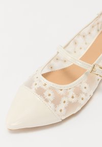 Chaussure plate blanche à bout pointu ; dotée d'un dessus en tulle transparent avec broderie de marguerites blanches et d'une fine bride de cheville avec des détails en or.