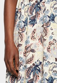 Blommönstrat tyg med stora blå och vinröda blad på en ljus bakgrund. En hand håller tyget nära fållen och visar dess textur.