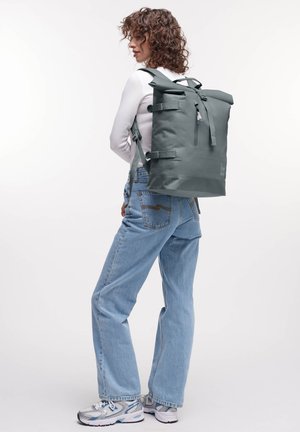Sac à dos gris à rabat, avec des sangles ajustables, une poche zippée et une texture lisse, imperméable. Associé à un jean bleu clair et des baskets.