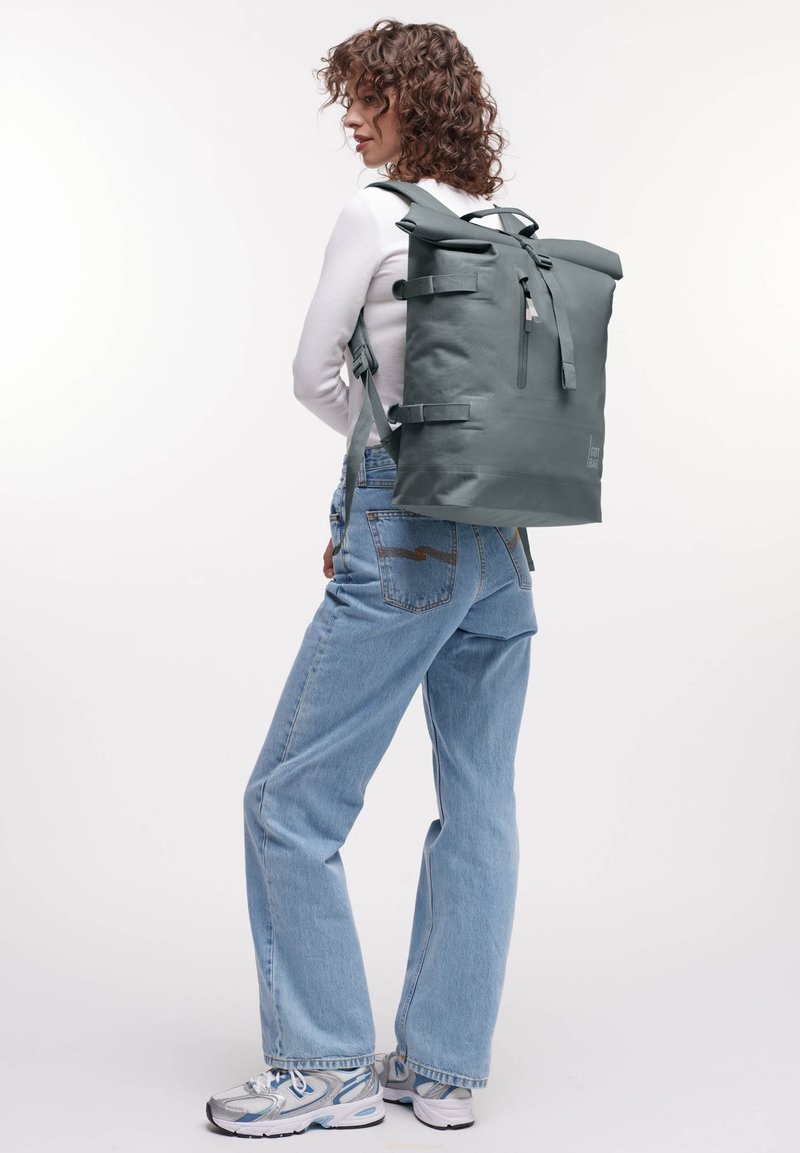 Zaino grigio a chiusura arrotolata con spalline regolabili, tasca con zip e una superficie liscia e impermeabile. Abbinato a jeans azzurri chiari e sneakers.