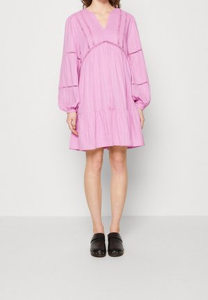 Frau trägt ein lockeres, knieumspielendes pinkfarbenes Kleid mit langen Puffärmeln und schwarzen Slip-On-Clogs mit genieteten Sohlen.