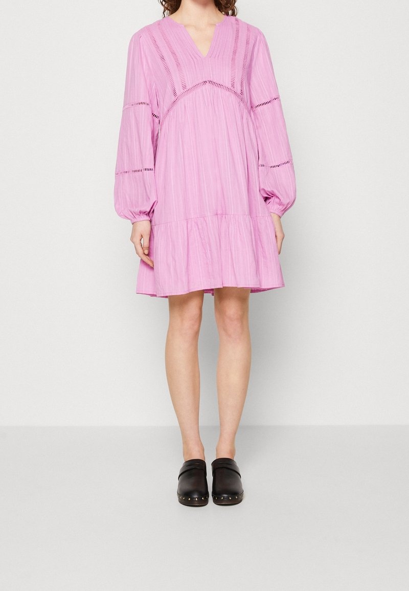 Femme portant une robe rose ample, longueur genou, à manches longues bouffantes, et des sabots noirs à enfiler avec des semelles cloutées.