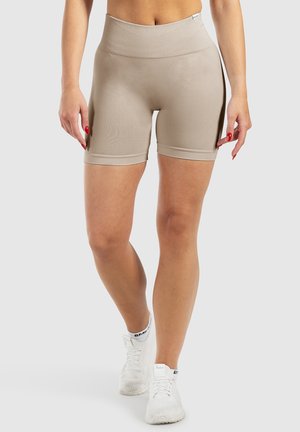 Femme portant un short cycliste beige taille haute et des chaussures de sport blanches, debout devant un fond clair uni.