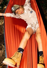 Enfant dans un hamac rouge portant un sweat-shirt beige, un short camouflage, des chaussettes noires et des bottes beiges. Le hamac a une texture lisse et une couleur vive.