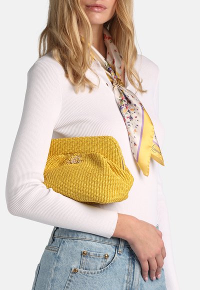 Femme portant un pull côtelé blanc et une écharpe fleurie, tenant une pochette tissée jaune et vêtue d'un jean bleu clair.