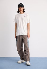Hvid t-shirt med et lille logo, beige bukser med blå accenter og hvide sneakers, båret med en sort kasket. Enkel og funktionel design.