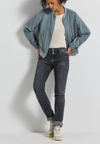Veste bomber bleu clair, t-shirt beige, jean foncé et baskets blanches ; avec fermeture éclair, poignets côtelés et jean retroussé.