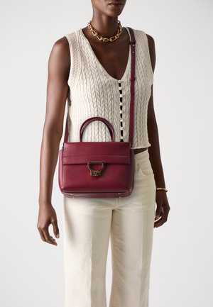 Burgundy leather handbag with dual handles, a detachable strap, textured finish, and gold hardware, styled against a neutral top and pants.