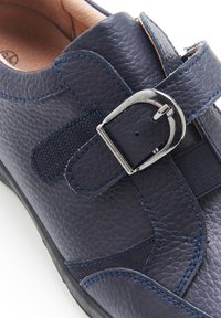 Chaussure en cuir marine avec une surface texturée, dotée de coutures bleues, d'une sangle avec une boucle en métal argenté, et d'accents élastiques pour un ajustement parfait.