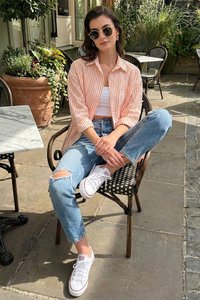 Woman wearing striped shirt, sunglasses, ripped jeans, and white sneakers sitting cross-legged on a chair in an outdoor café with plants nearby.