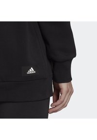 Czarna bluza z okrągłym dekoltem, z prążkowanymi mankietami i dół. Z boku widoczna mała, biała naszywka z logo Adidas. Materiał wydaje się miękki i teksturowany.