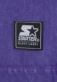 Violetinės medžiagos pleistras su baltu logotipu "STARTER® BLACK LABEL", apsuptu juodo krašto, rodančiu tekstūruotą mezginio raštą.