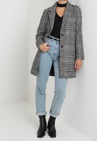 Kvinna i svart topp, grå rutad kappa, ljusblå jeans med uppvikta ben och svarta ankelskor som står mot en vit bakgrund.