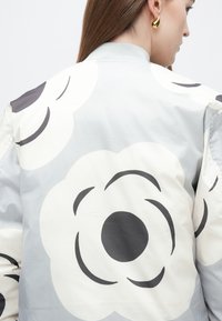 Lekka kurtka bomberka w pastelowo szarym kolorze z dużymi wzorami kwiatowymi w kolorze białym i ciemnoszarym; posiada prążkowany kołnierz i gładką fakturę.