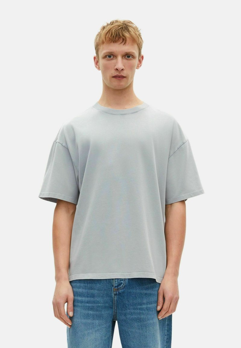 Camiseta de corte amplio en gris claro, hecha de algodón, con cuello redondo, hombros caídos y mangas cortas, combinada con jeans azules.