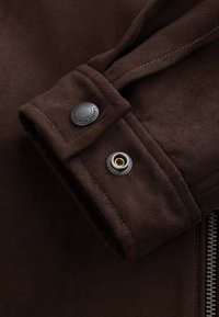 Dark brown suede jacket sleeve with stitched cuff and metal snap button fastener, part of a visible zipper on right side.