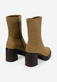 Un par de botas de gamuza color tostado hasta la mitad de la pantorrilla, con tacones de bloque apilados en marrón oscuro y cremalleras laterales, mostradas desde la parte trasera y ligeramente anguladas.