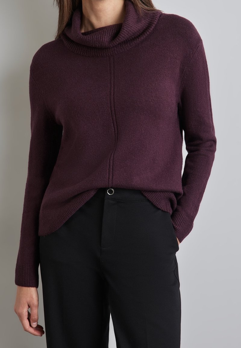Pull oversize violet foncé avec un col bénitier, des détails côtelés et une coupe décontractée, associé à un pantalon noir élégant avec un bouton.