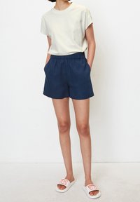 Světle šedé tričko s krátkým rukávem a volným střihem, doplněné o tmavě modré kraťasy s elastickým pasem a bočními kapsami. Bílé pantofle s červeným potiskem.