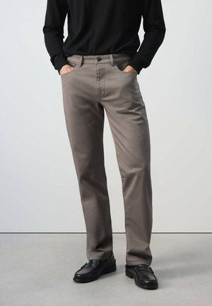 Persona que lleva pantalones estilo taupe de pierna recta, camiseta negra de manga larga metida dentro y mocasines negros, de pie con las manos en los bolsillos.