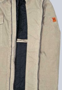 Beige Cordjacke aus Kord mit schwarzem, gestepptem Innenfutter. Verfügt über einen Frontreißverschluss und einen orangefarbenen Logo-Patch auf der oberen linken Seite.