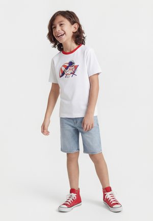 Αγόρι που χαμογελάει και στέκεται, φοράει λευκό μπλουζάκι με γραφικό αστροναύτη, ανοιχτόχρωμα τζιν σορτς και κόκκινα sneakers ψηλού τύπου.