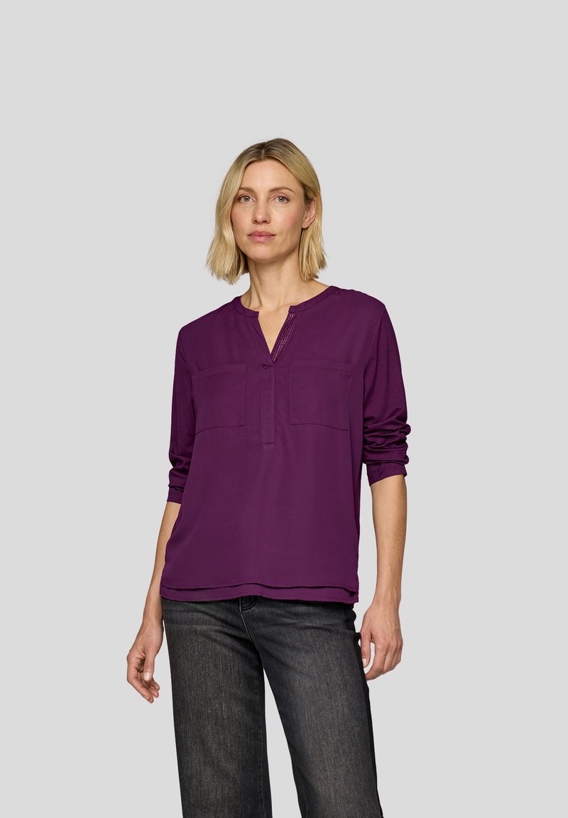 Lila Bluse mit V-Ausschnitt, zwei aufgesetzten Taschen vorne und langen Ärmeln. Weicher, leichter Stoff mit glatter Textur und entspanntem Schnitt.