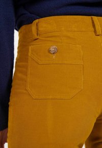 Pantalons en velours côtelé jaune avec une poche arrière dotée d'une fermeture à bouton. Le tissu présente une texture côtelée, avec des accents de couture visibles le long des coutures.