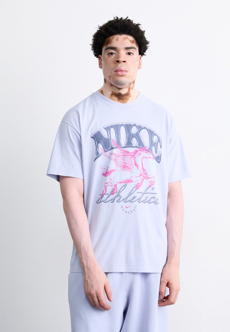 Lyseblå bomulds t-shirt med et pink vingeret heste grafik og "NIKE athletics" tekst. Enkel design med korte ærmer og løs pasform.