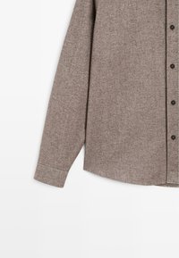 Brun flannel skjorte med knap-front, lange ærmer og en buet kant. Tekstureret materiale med sorte knapper langs stolpen.