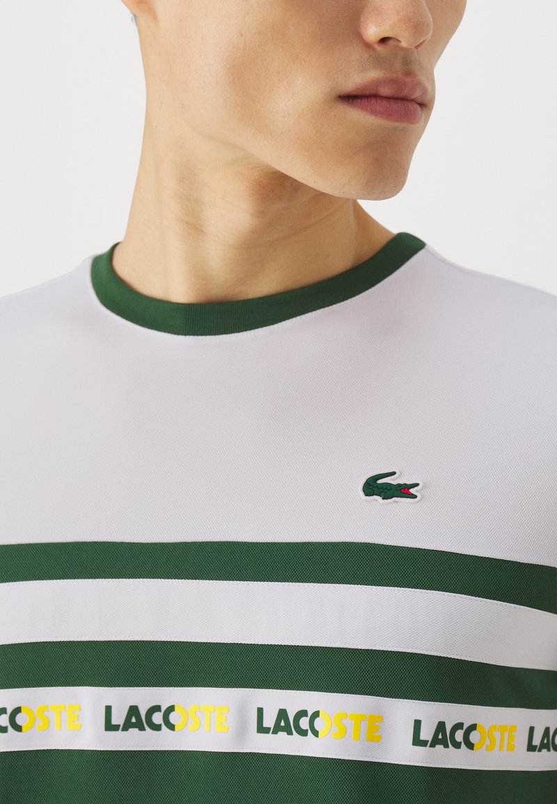 Polo blanc à manches courtes avec des accents verts, présentant des rayures horizontales et un petit logo de crocodile vert. Fabriqué en matériau texturé.