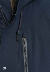 Veste bleu marine en gros plan montrant une poche avant zippée, une fermeture éclair et un cordon réglable avec un curseur sur tissu texturé.