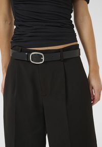 Zwarte leren riem met een vierkante zilveren gesp, gedragen over hoge zwarte broeken. Beschikt over een gladde textuur en een klassiek ontwerp.