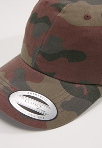 Cappello da baseball mimetico in tonalità di verde e marrone, con visiera curvata, tessuto testurizzato e un adesivo con marchio argentato sulla parte frontale.